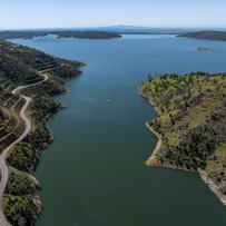 An aerial view of Lake Oroville with the Bidwell Canyon marina in the background and a winding road and hillside to the left. 