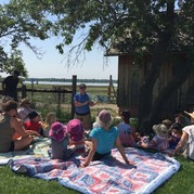 A group of children sit on a blanket under the sun as a volunteer reads a book.