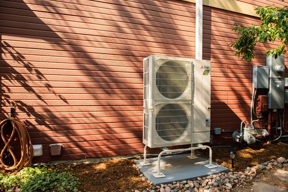 A white heat pump condenser unit stands on a platform outside a house with red siding. 