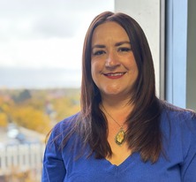 Christa wearing a blue sweater and standing in front of a window with trees in the distance