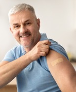 Man with gray hair smiles as he holds up his sleeve to show a bandage on his arm
