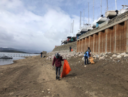 People stroll along the shore, carrying bright orange bags against a backdrop of sand and sea.
