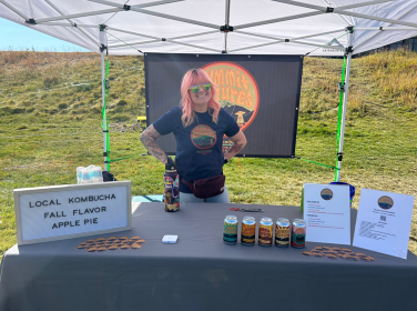  A woman with pink hair stands behind a table displaying various bottles of kombucha.