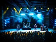 A band performs on stage, illuminated by vibrant lights, creating an energetic atmosphere for the audience.