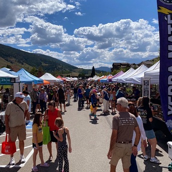 A bustling outdoor market scene with a diverse crowd and colorful tents set up for vendors and shoppers.