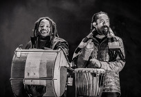 Two men with dreadlocks stand side by side, each holding a drum, showcasing their musical connection.