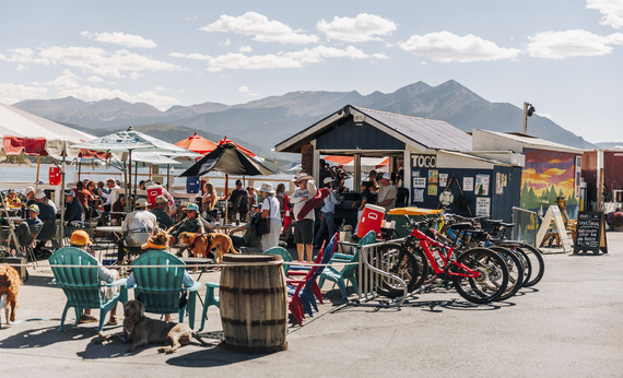 A group of people relaxes at outdoor tables, accompanied by their bikes and dogs, in a vibrant outdoor atmosphere