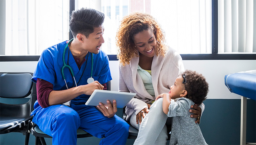 Smiling health care professional talks to a mother and her young daughter in a hospital lobby.