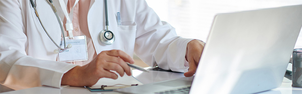 A doctor works at a laptop in a medical office setting.
