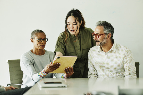 Two people sitting looking at a tablet while another person stands and points to the tablet