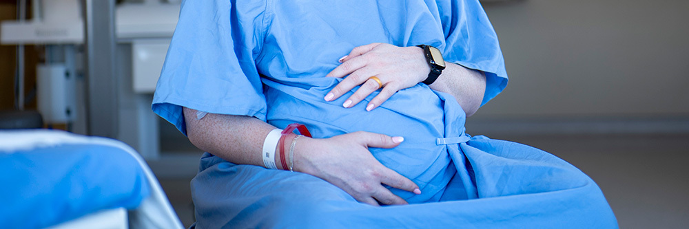 A pregnant woman in a hospital gown holds her hands over her abdomen.