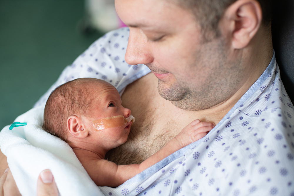 A new father cradles his premature baby between his hospital gown and his bare chest in an example of "kangaroo care".