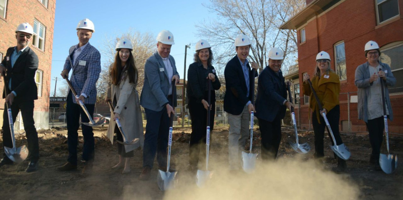Nine people wearing white hardhats and scooping with shovels at the groundbreaking event.