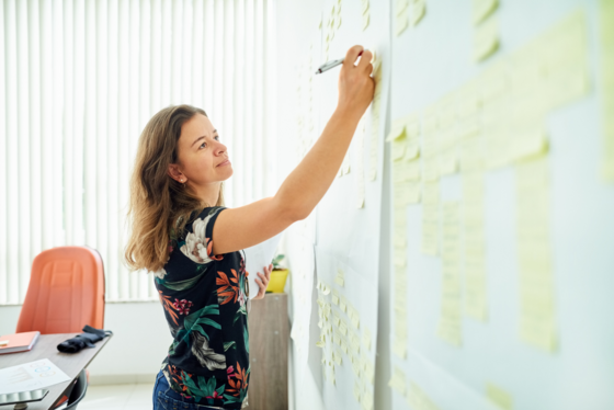 A woman stands in front of a whiteboard with sticky notes with a pen in hand. 