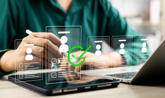 Person reviewing digital candidate profiles on a tablet and laptop, with a green check mark indicating selection.