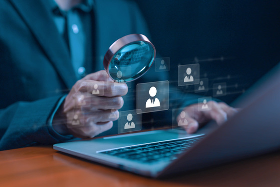 Person using a magnifying glass to examine digital user icons on a laptop, representing recruitment or talent search.