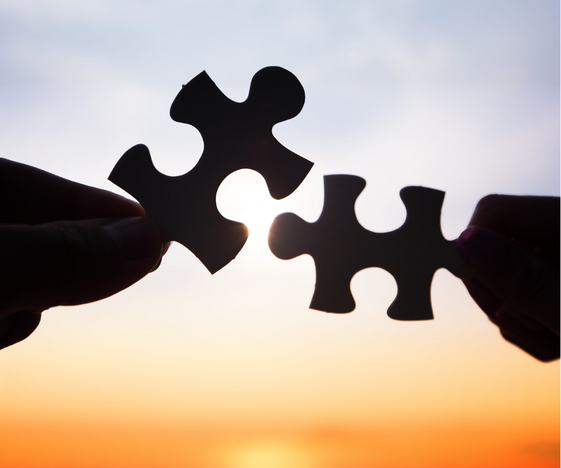 Two hands hold puzzle pieces toward each other in front of a sunset, symbolizing connection, teamwork, or collaboration.