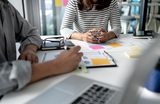 Two people sitting at a desk reviewing documents and charts with colorful sticky notes, pens, a laptop, and glasses on the table.