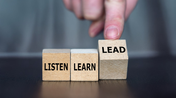 Three wooden blocks read “Listen,” “Learn,” and “Lead,” with a hand pushing the “Lead” block forward.