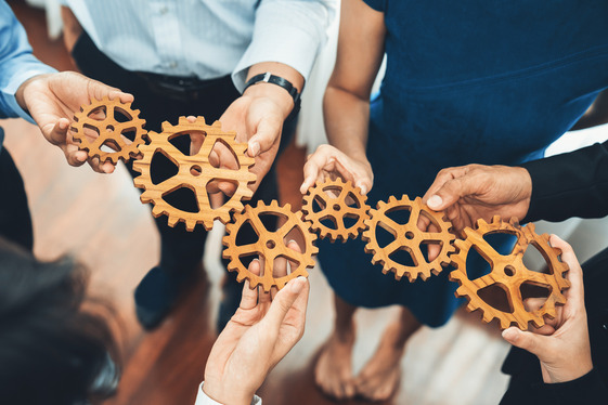 Group of people holding interlocking wooden gears, symbolizing teamwork and collaboration.