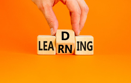 Hand adds a wooden ‘D’ block to blocks spelling LEARNING—visual for L&D (Learning & Development) on an orange background.