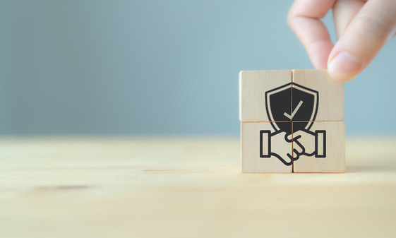 Hand stacking wooden blocks with a handshake and shield icon symbolizing trust and partnership.