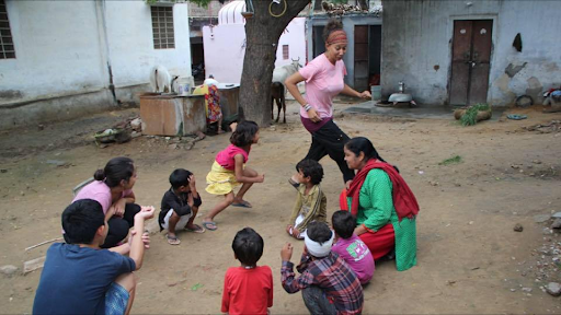 Rosina interacts with a group of children in an outdoor courtyard; some sit while others stand or jump, smiling and engaged in the activity.