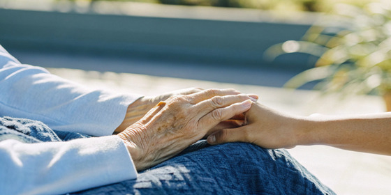 A younger person gently holds the hand of an older adult, conveying comfort and support in an outdoor setting.