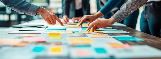 Hands of several people reach across a table covered with colorful sticky notes and papers during a collaborative planning session.