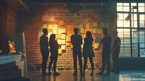 A group of people stand in front of a wall covered with colorful notes, discussing ideas in a warm, collaborative workspace.