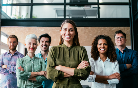 A diverse group of six professionals standing together in an office, all smiling with arms crossed, with a woman in an olive shirt centered in front.