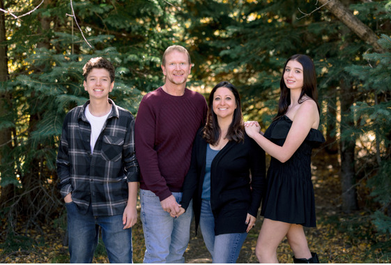 Four people standing together outdoors in front of evergreen trees, smiling at the camera.