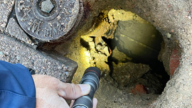 A worker shines a flashlight down a hole to reveal a water service line.