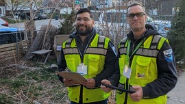 Two men wearing yellow vests and holding clipboards stand in a yard.