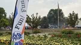 A flag reading "Fun! Fun!" waves in the breeze in front of a stone Lincoln Park sign surrounded by yellow flowers.