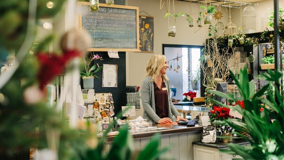 A blonde woman laughs at the check-out counter of a plant shop full of greenery and vines