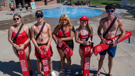 Lifeguards pose in front of Discovery Bay Waterpark splash pad with family in the background