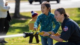 A female firefighter helps a young girl spray a fire hose while they both smile and laugh on a sunny fall day.