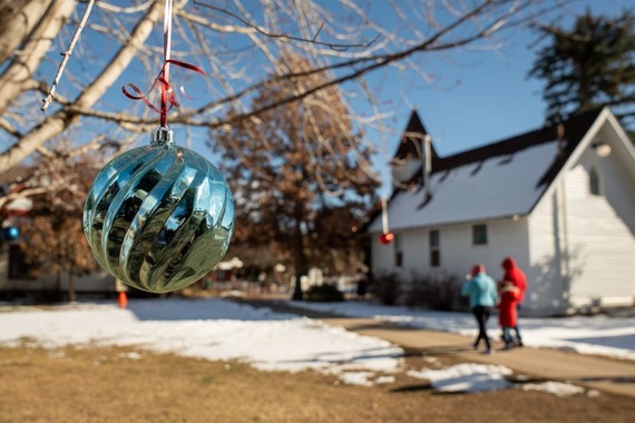 A metallic blue ornament hangs from a snowy tree outdoors near a white clapboard one-room church at Centennial Village