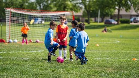 Young girls and boys play soccer in the grass, with a quick huddle, while a girl in goal watches