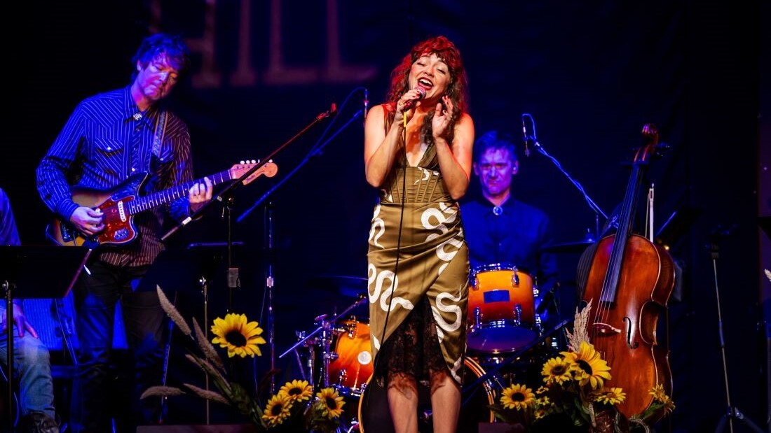 A woman sings under a warm spotlight in front of her band. The floor is covered in sunflowers, and there are snakes on her dress.