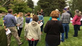 A man speaks to a standing crowd on a grassy lawn surrounded by trees