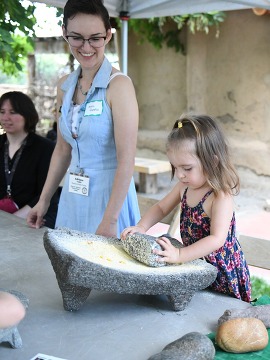 little girl uses mano y metate to grind corn while an adult looks on