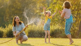 A woman sprays two laughing children with a sprinkler in the yard.