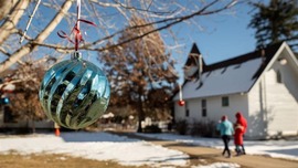 A teal ornament hangs from a tree branch. A family walks nearby a church with snow on the ground in the background.