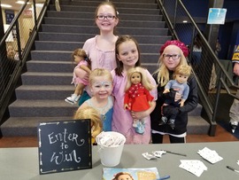 Four young children smile while standing behind a table holding American Girl Dolls. Stairs at the Greeley Ice Haus are in the background.