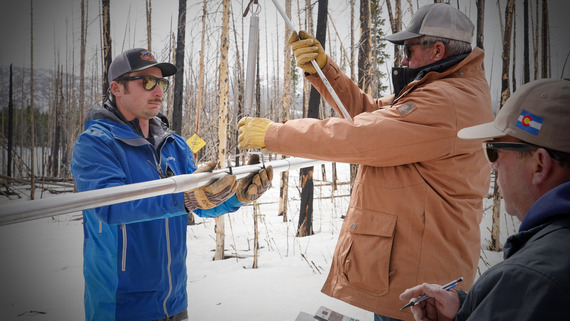 Two men weigh a long pipe containing snow while another waits to do calculations.