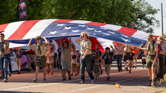 Boy Scouts carry a large flag over their heads while walking.