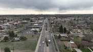 Aerial view of a suburban road with houses, cars, and cloudy sky, city skyline visible in the distant background.