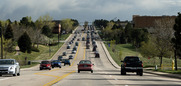 Cars drive in both directions on a wide, tree-lined road under a cloudy sky, with light traffic and green grass.